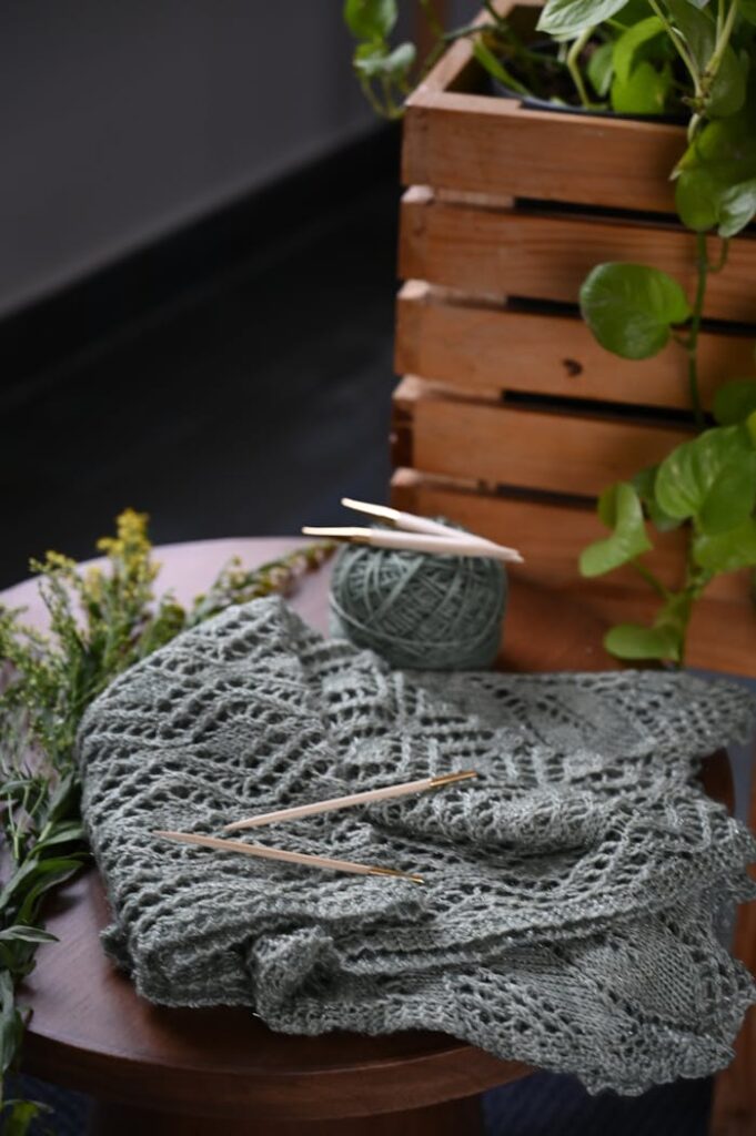 Close-up of a crochet project with yarn and needles, surrounded by greenery.